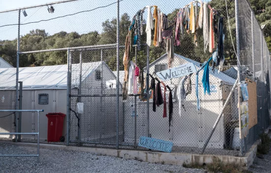 A refugee camp entrance is pictured with a large wired fence surrounding it. Hand made welcome signs and a heart made of old clothes are visible.