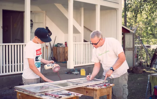 A younger and older man are seen together working to fix an old window frame. The house being fixed is visible in the background.