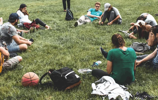 A group of young people are sitting on grass and talking in a circle formation. Bags and sports gear can be seen lying around them.