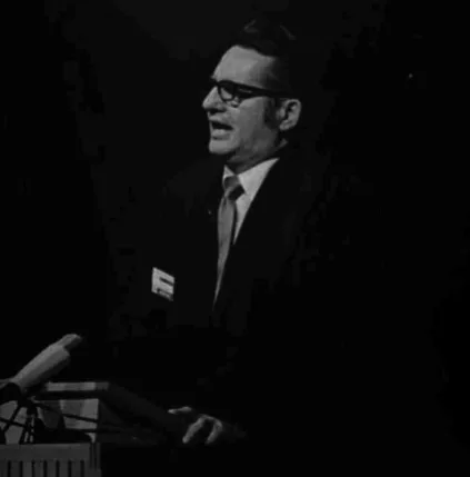 Bob Evans The founder of GEM in an older black and white photo. He is wearing a black suit and is in the midst of speaking into a microphone whilst looking off camera.